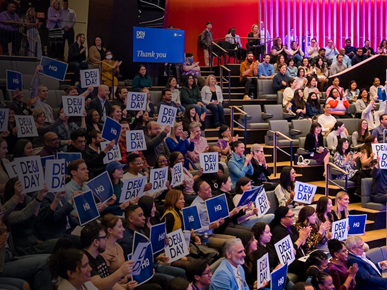 Crowd sitting in a theater at DE&I Day holding signs