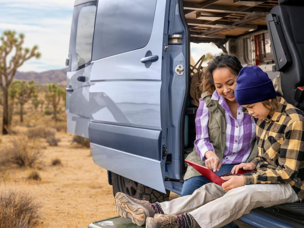 An adult and hild looking at an electronic tablet while sitting in the open side of a van in an arid area.