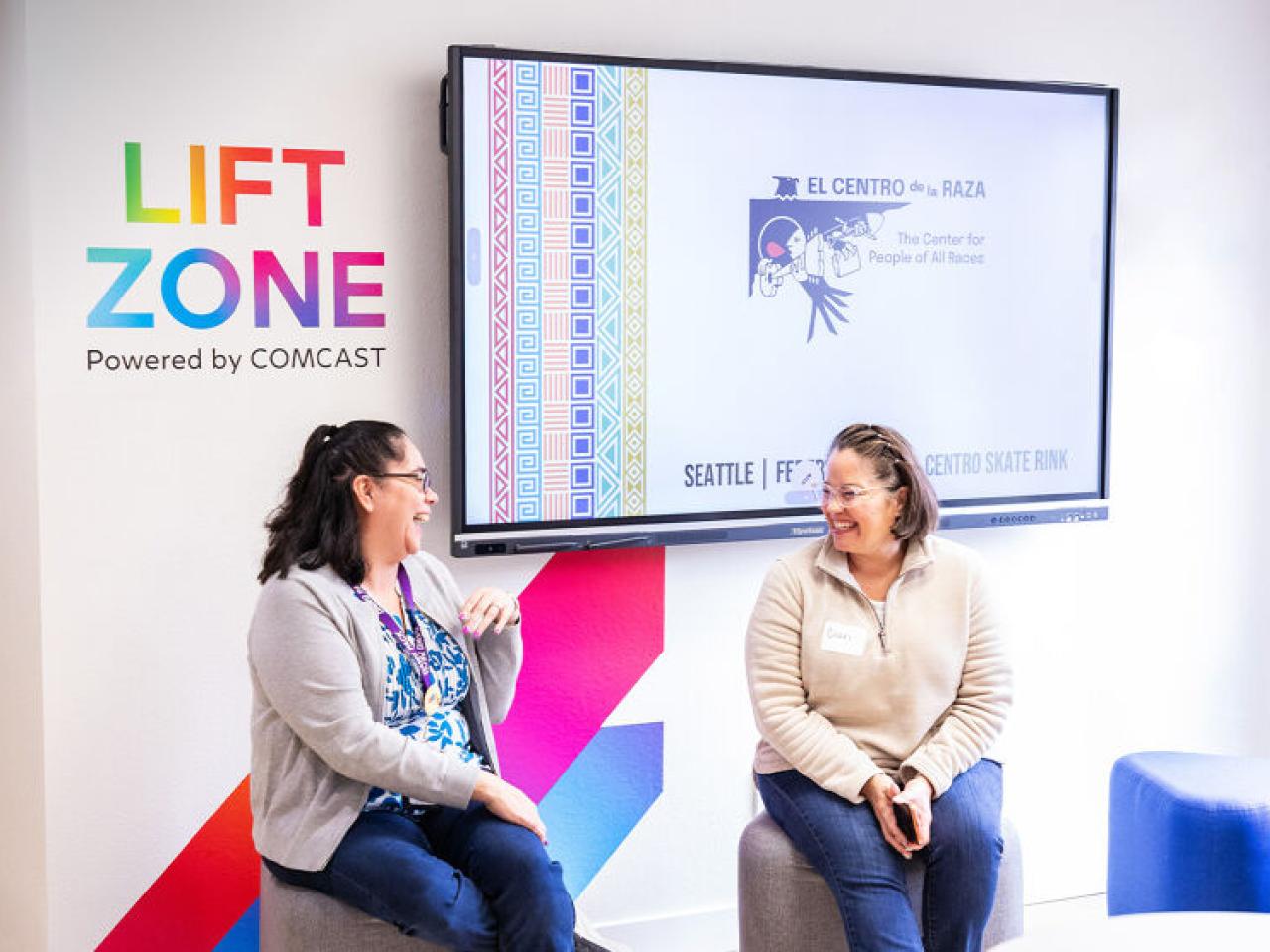 Two people sitting and talking in front of a mural that reads "Lift Zone, Powered by COMCAST"