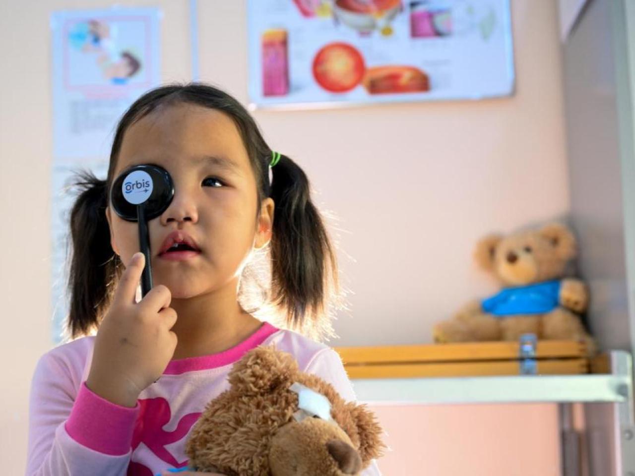A child holding a tool to cover one eye. Holding a teddy bear with the other hand.