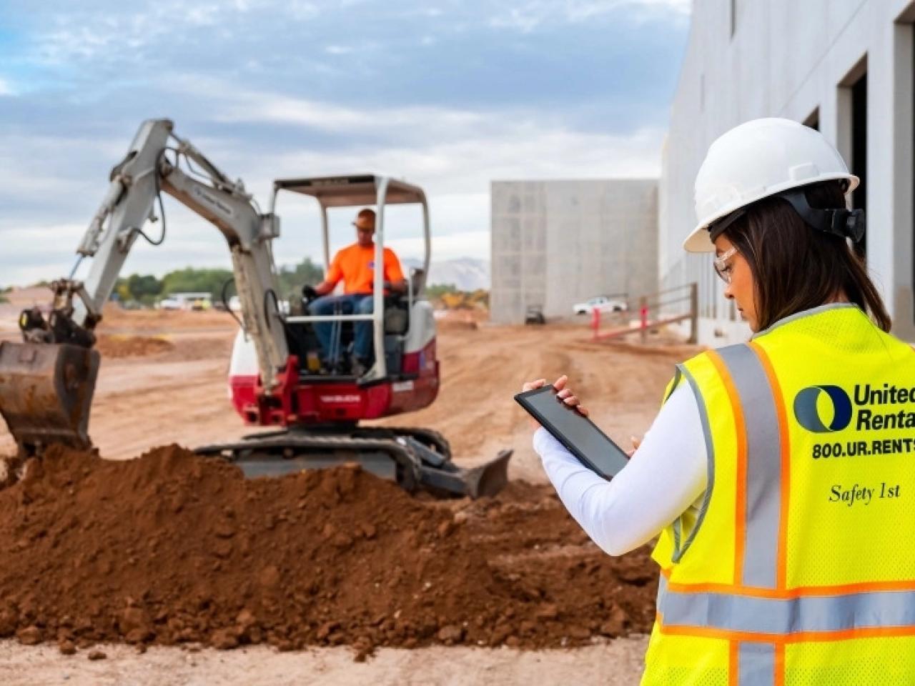 A person looking at a tablet. Another in the distance using a large digging machine.