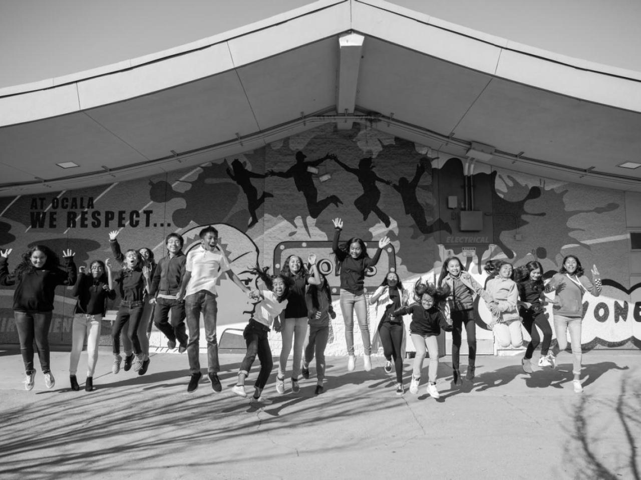 Black and white photo of a group of kids in a line jumping. A covered stage behind them.
