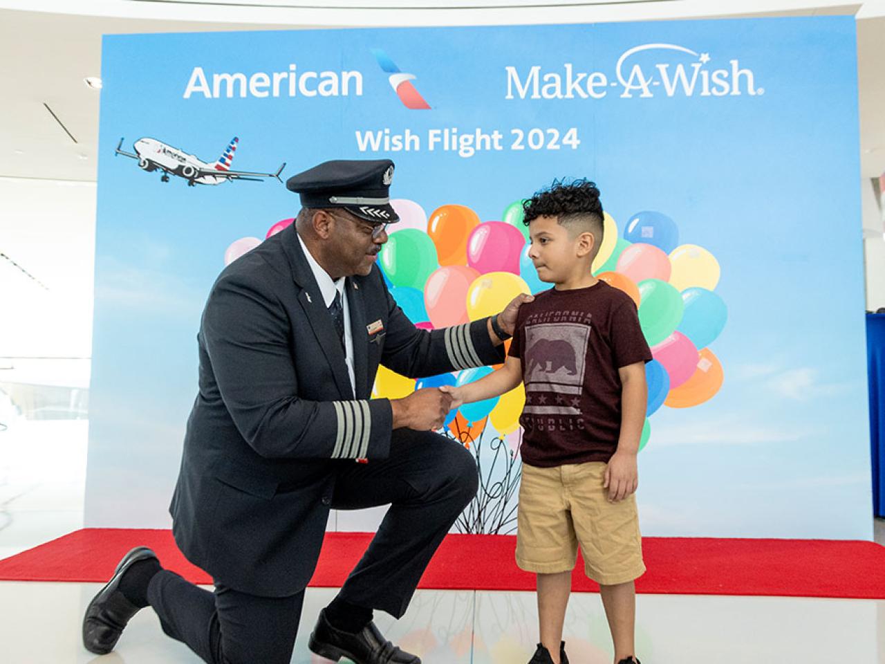 A pilot kneels and shakes the hand of a child. A poster behind them with American and Make a Wish logos, balloons.