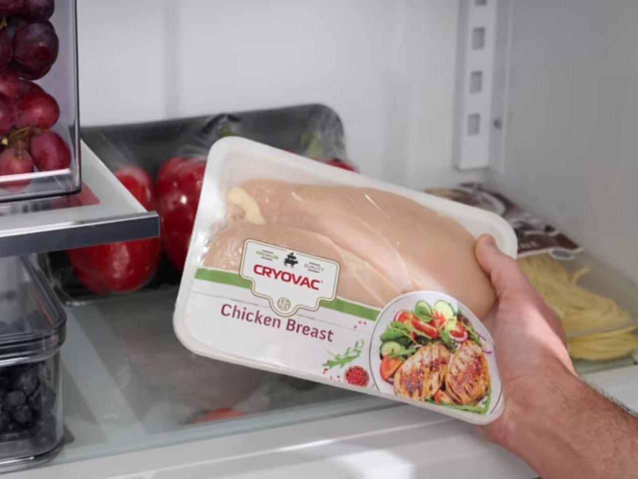 A hand holding a package of "Chicken Breast" in a refrigerator.