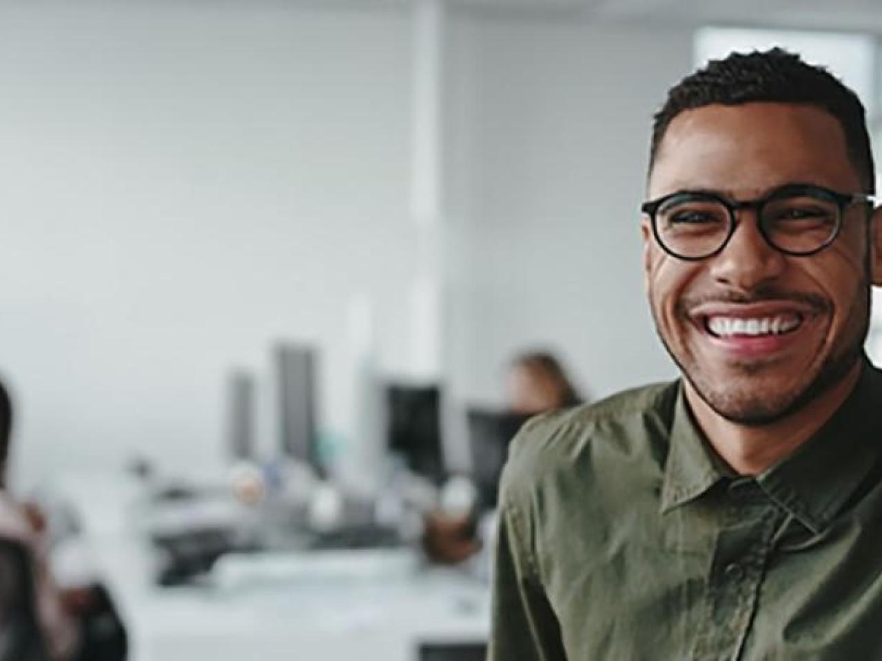 A smiling person looking at the camera, a small group of people at a shared desk behind them, in an office setting