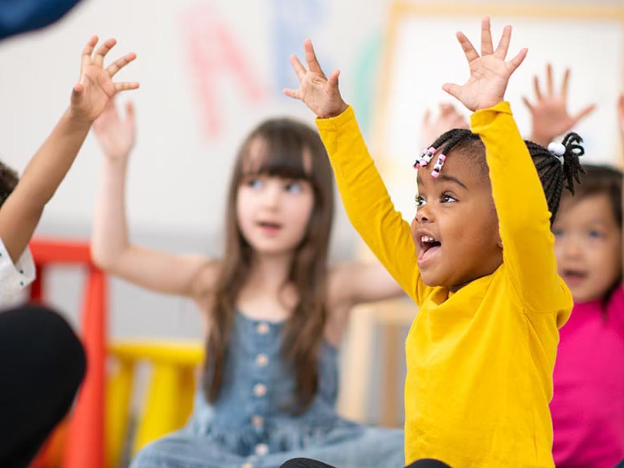 Children seated in a classroom setting, all with raised hands.