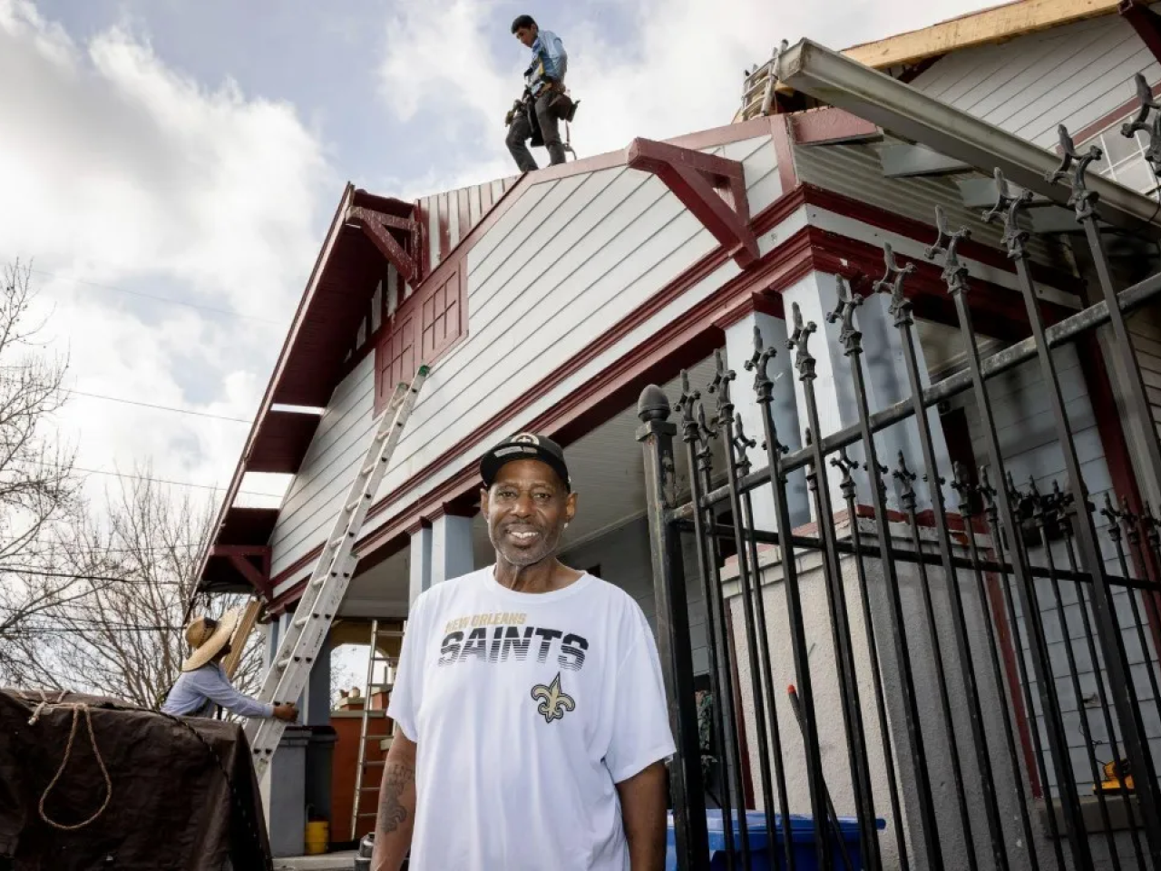 A person in a New Orleans Saints t-shirt standing in front of a house with people doing work on it