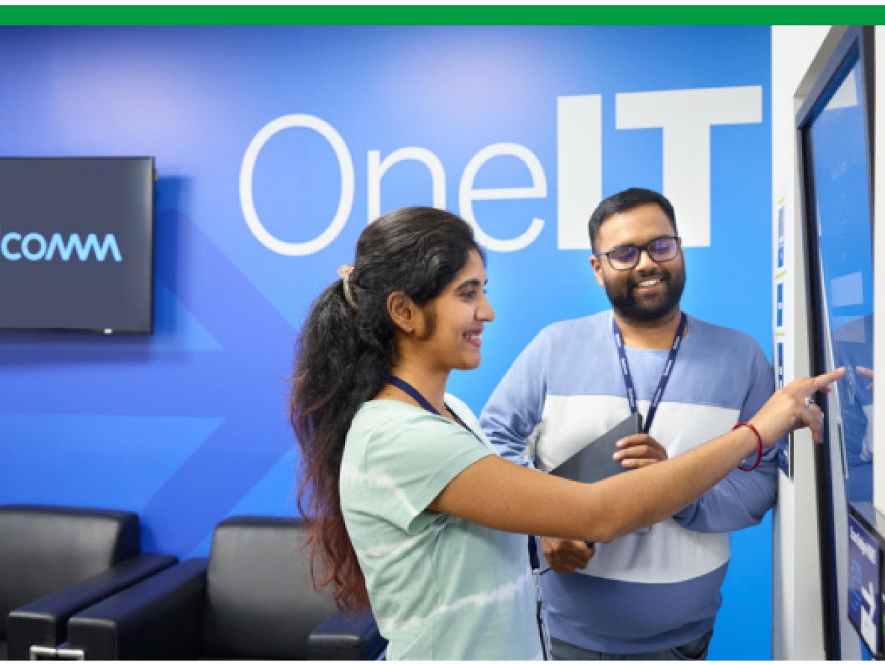 Two smiling people looking at a large touch-screen display. "OneIT" on the wall behind them.