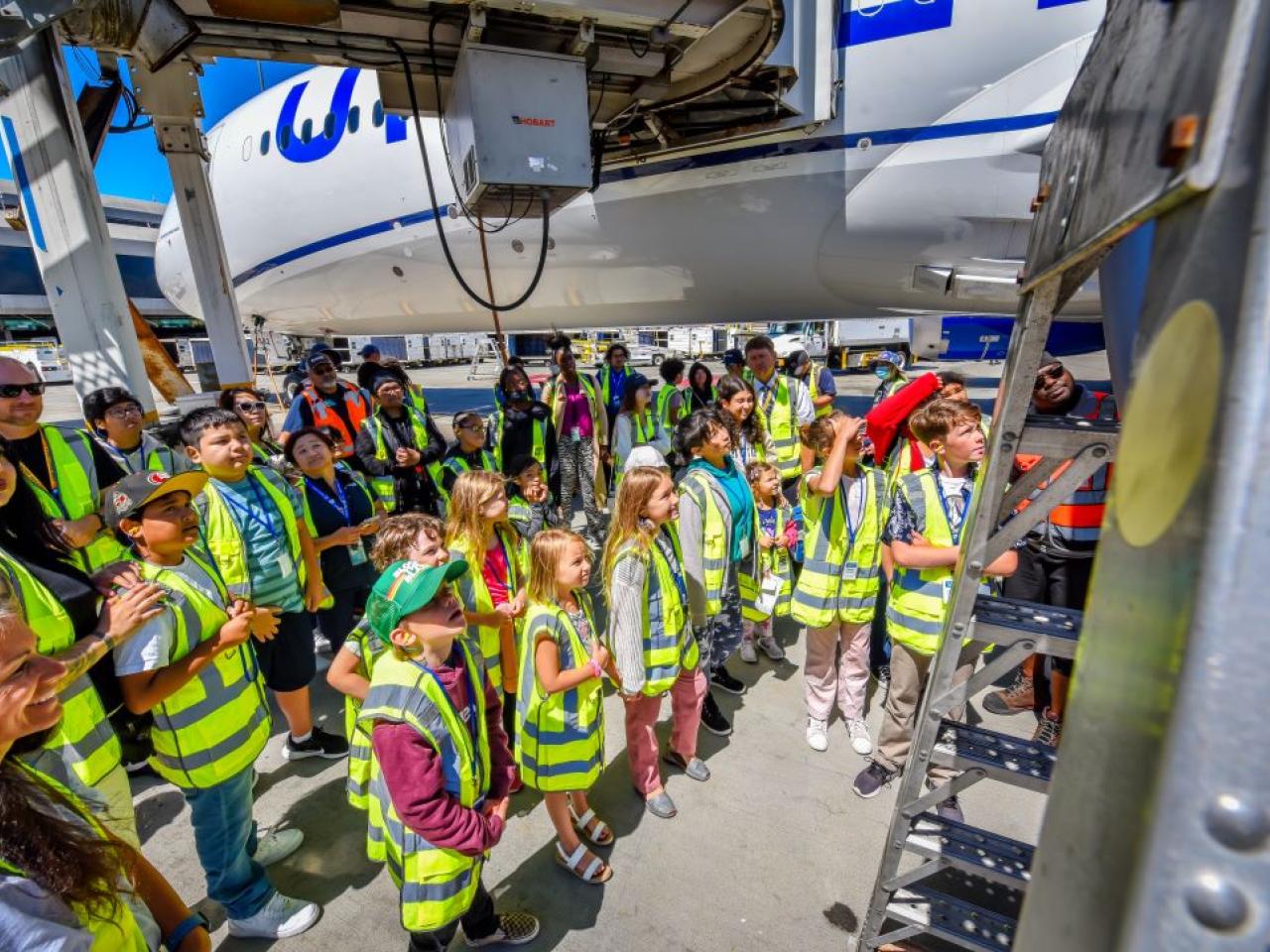 A group of kids and adults in high-vis vests looking up close at an airplane.