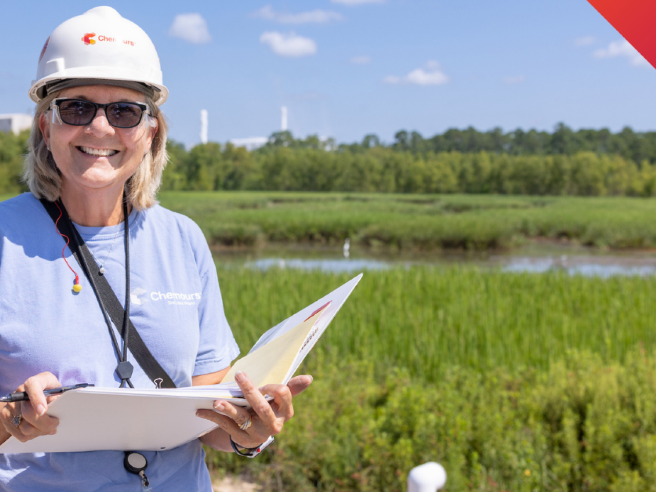 A person smiling outside, holding a notebook and wearing a hard hat
