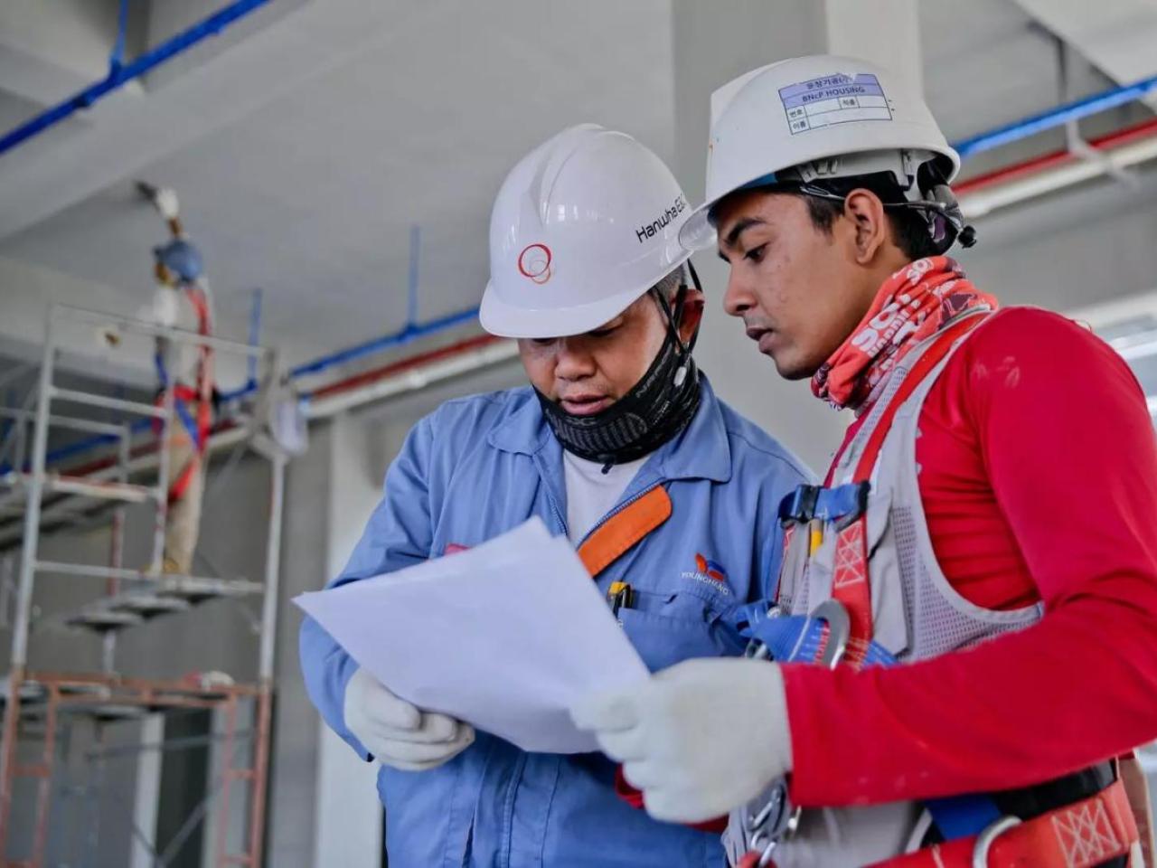 Two people in hard hats, gloves and protective masks looking at a piece of paper. A person standing on scaffolding painting the ceiling behind them.