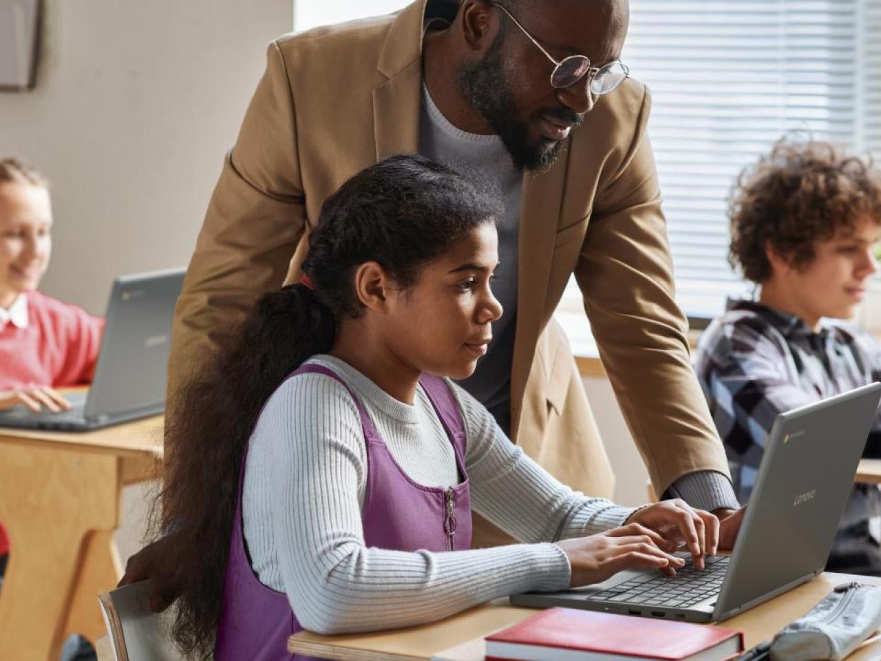 A teacher helping a student in a classroom, using a laptop