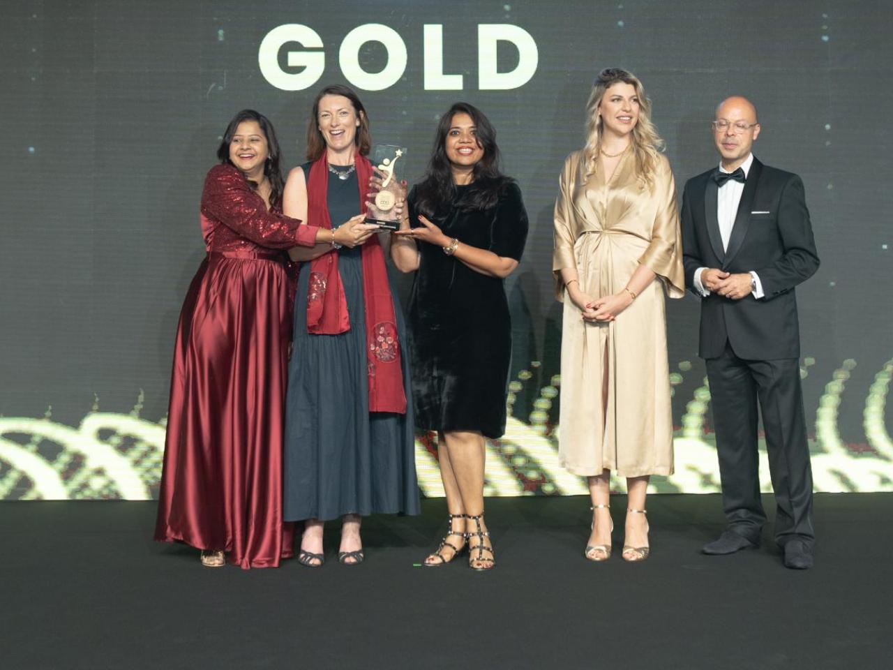 Five people on a stage in front of the word "GOLD", three holding an award