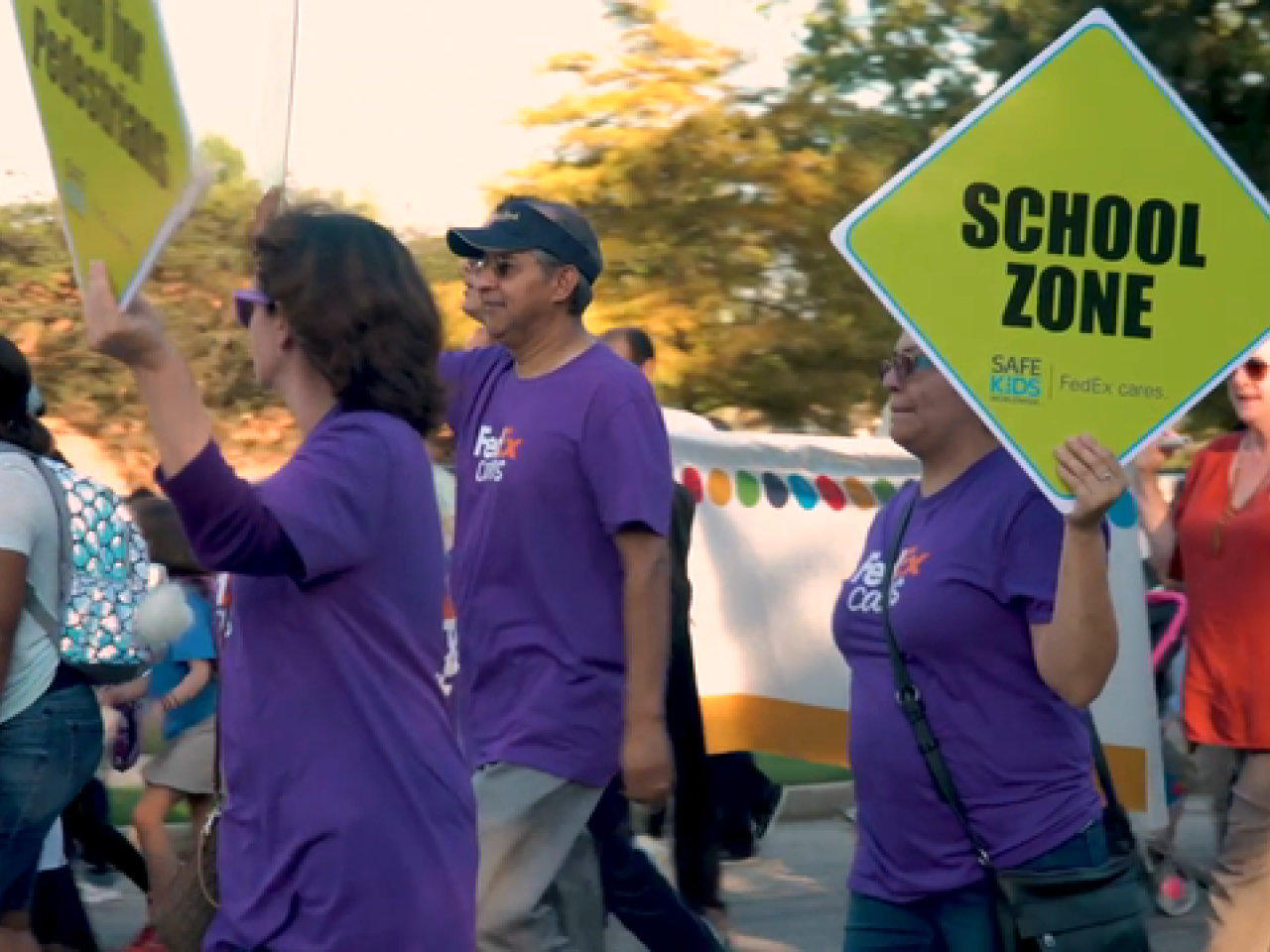 3 people in purple FedEx shirt, 2 of which are holding neon yellow "School Zone" signs