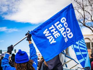 Person wearing blue holds a blue flag with the words, "good leads the way" printed in white on the flag