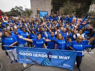 A large group of people wearing blue stand behind a blue parade banner with the words, "good leads the way" written in white