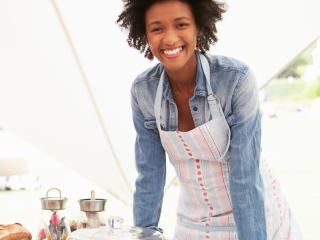 a person wearing an apron in a white tent, baked goods in front of them
