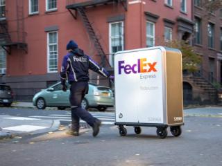A person in FedEx uniform pulling a tall cart with FedEx Express logo on it crossing a street.