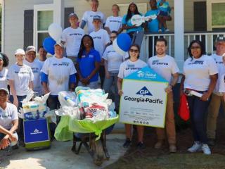 A group of volunteers in front of a house. All wearing shirts that say "GP United". A wheelbarrow full of supplies in front of them and a Georgia-Pacific sign being held by two.