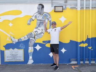 LA Galaxy player Landon Donovan poses with his arms outreached in front of a yellow, blue and white mural of him in a LA Galaxy jersey kicking a soccer ball. 