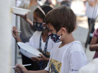Elementary students wearing masks, each hold bowls of paint and brushes as they stand in a line and finish the LA Galaxy mural at their school. 