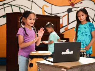 children standing in front of a laptop, one behind them playing on a piano. Wall behind them with a painted mural of floating music notation.