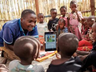 A group of children in a grass-walled hut, an adult showing them an electronic tablet with a drawing on it.
