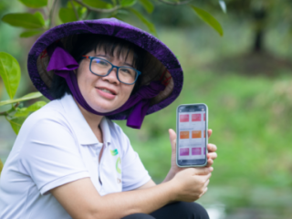 Nguyen Thi Kim Thoa sitting in an orchard showing a cellphone to the camera. She is wearing a large rimmed hat.