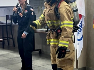 A person dressed in full fire-fighting PPE and a person next to them with a microphone.