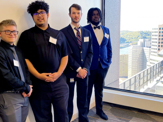 Five students in business style clothing standing in a hallway with a large window at the end overlooking a city landscape.