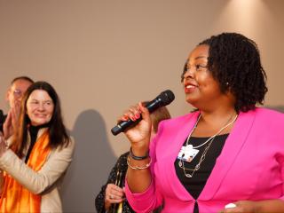 Salah Goss with a microphone, others to her side clapping and smiling.