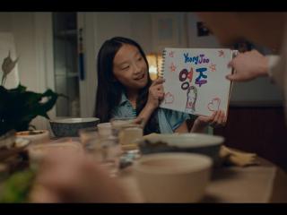 A child at a dinner table holding a notebook with hand-colored symbols, pictures and the name "Yeong Joo." An adult points to it.