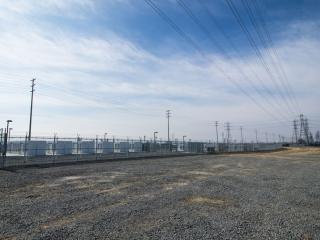  A power station with tall power lines above and graveled pad in front.