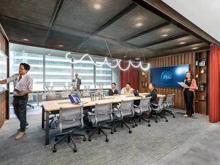 People seated at a long table with rolling office chairs, a long open wall, and person at a board near the front of the space.