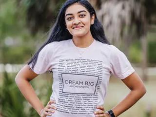 A person smiling at the camera, hands on their hips. Wearing a "dream big" t-shirt. 