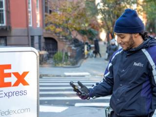 A person in FedEx uniform next to a tall cart with FedEx Express logo on it. They are looking at a hand-held device on a sidewalk in a residential area.