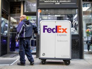 A person in FedEx uniform pulling a tall cart with FedEx Express logo on it, waiting outside of a jewelry store.