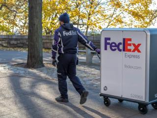 A person in FedEx uniform pulling a tall cart with FedEx Express logo on it through a park setting.