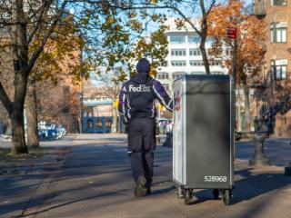 A person in FedEx uniform pulling a tall cart with FedEx Express logo on it down a sidewalk as a car goes by.