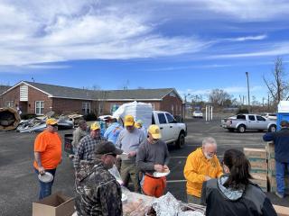 Line of people waiting to be served food in a parking lot. Some storm damage seen behind them at a nearby building.