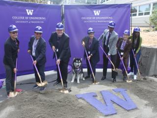 seven people in hard hats holding shovels dug in to sand around a large "W" and one very good dog in the middle