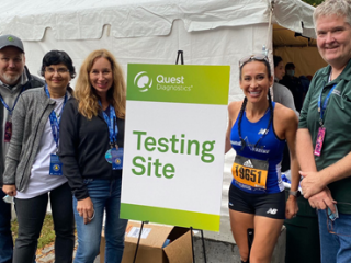 a group of five people, one in running apparel, by a sign for Quest "Testing Site" and a white tent behind them