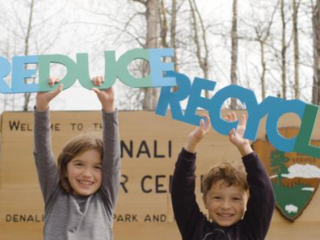 Two children stand in front of a Denali park sign holding up words "Reduce" and "Recycle"