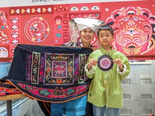 a child stands beside Mrs. Huang who is holding an embroidered sling. Cultural art behind them