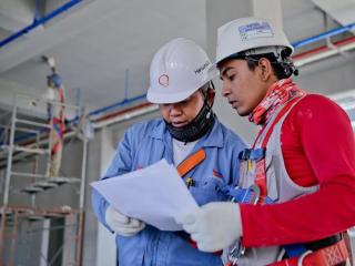 Two people in hard hats, gloves and protective masks looking at a piece of paper. A person standing on scaffolding painting the ceiling behind them.