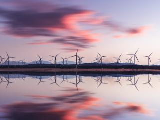 A group of wind turbines behind a body of water. The setting sun to the right.