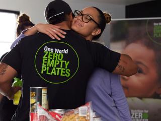 A person hugging someone with a t-shirt that reads: "One World, Zero Empty Plates"