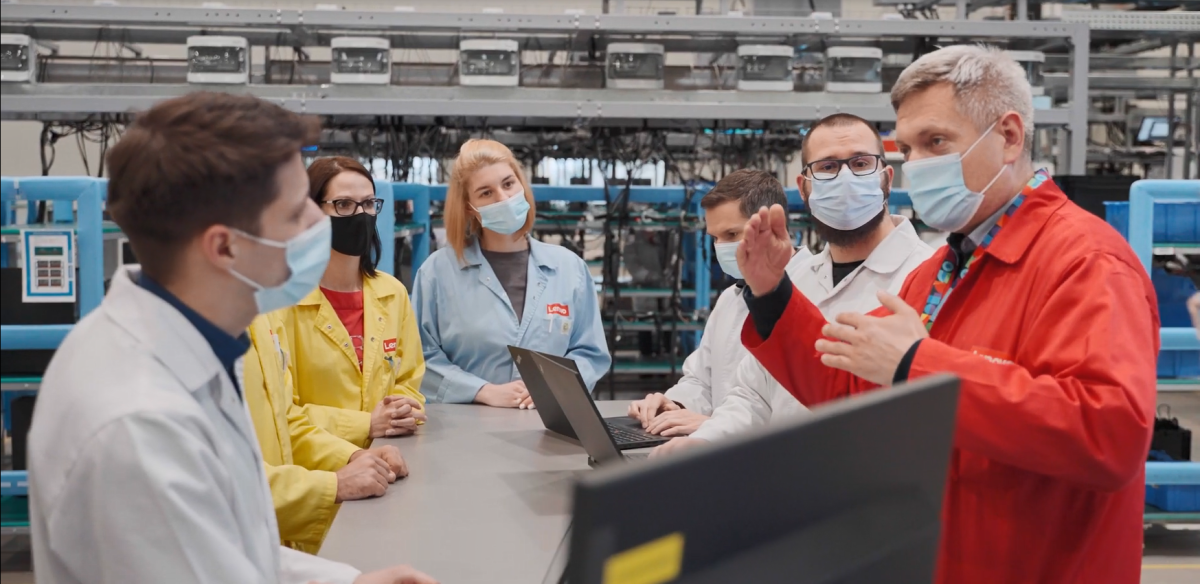 small group of people gathered around a table, all wearing protective masks and lab coats of different colors. Laptops open on the table. Factory machinery behind them.