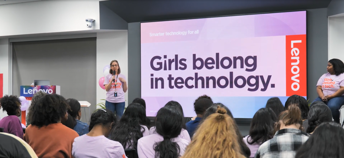 A person on stage with a microphone. "Girls belong in tech" on a large screen behind them.