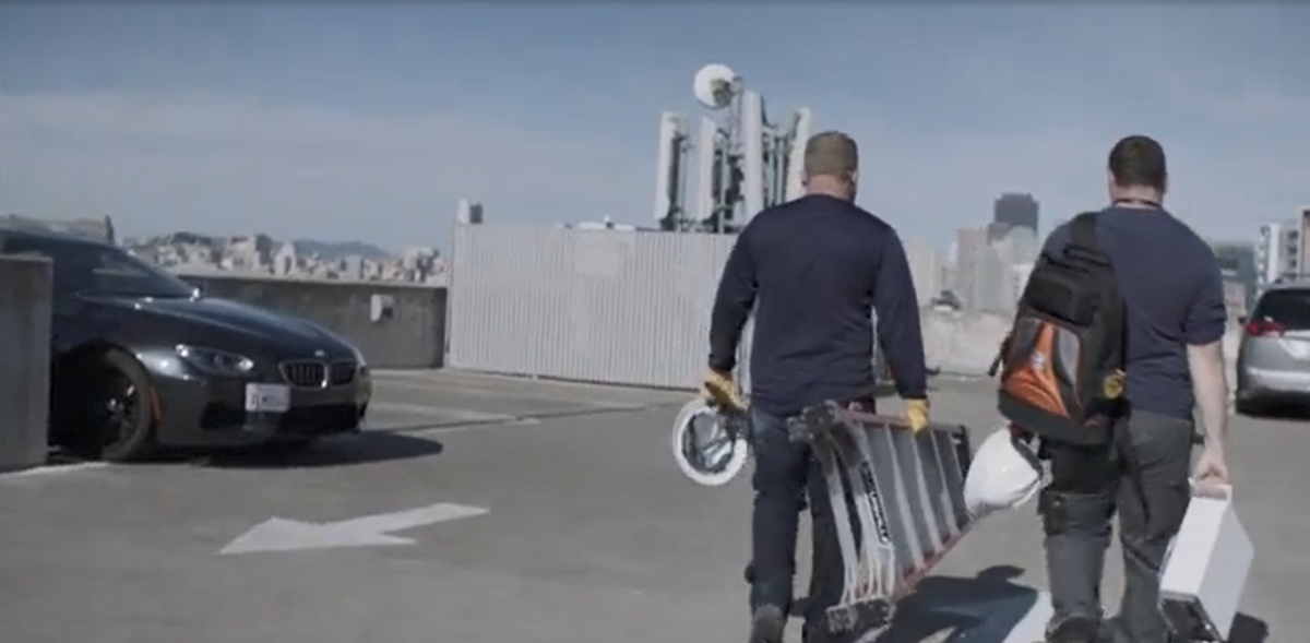 Two workers walking on a roof-top parking structure. One carries a ladder and hard hat,  the other carries a hard hat and white box.